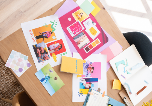 Wooden table with a variety of colorful postcards, a book, a pen, a flower vase with yellow flowers
